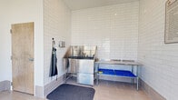 This single-floor Pet Spaw is a purpose-built grooming facility designed to make pet care clean, comfortable, and efficient. The space is anchored by a professional-grade stainless steel wash tub equipped with integrated fixtures and a splashguard, accompanied by a stainless steel prep and drying table with lower storage. White subway tile lines the walls from floor to ceiling, creating a bright, hygienic atmosphere that is easy to maintain. Large-format neutral floor tiles add to the clean, polished aesthetic throughout.

Near the entrance, a floor-to-ceiling glass door and oversized window flood the space with natural light while offering views of lush landscaping and palm trees outside. A sanitation station with a stainless steel wipe dispenser is conveniently positioned at the entry point. Wall-mounted soap and product dispensers, along with hooks for aprons and accessories, keep grooming essentials within easy reach. Posted guidelines ensure the facility is used responsibly and kept clean for all users.

A wood-tone interior door provides access to adjoining areas, and pet-themed artwork adds a welcoming, playful touch to the otherwise functional space. The overall design balances practicality with a polished, modern finish, making it a well-equipped amenity for pet owners.