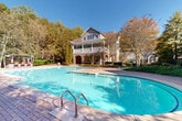 Located at 100 Ardsley Pl in Norcross, Georgia, this outdoor amenity space centers around a generously sized resort-style swimming pool set against a picturesque backdrop of mature trees. The pool area features a classic kidney-curved design with a separate wading section, surrounded by a well-maintained brick paver deck that provides ample space for sunbathing and relaxation. A prominent white pergola runs along one side of the pool, offering shaded seating beneath its open-beam structure and lined with lounge chairs for residents to enjoy. An additional covered cabana area with casual seating is positioned nearby, adding to the social atmosphere of the space. The surrounding landscape is densely wooded, providing natural privacy and a serene, park-like ambiance throughout the seasons. A multi-story clubhouse building anchors one end of the pool deck, featuring stone and siding exterior details, large windows, and a covered upper balcony that overlooks the pool. The architecture blends traditional residential design with comfortable outdoor living. The entire area is enclosed with fencing and benefits from abundant natural sunlight, making it a well-appointed community gathering space ideal for leisure and recreation.