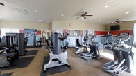 This single-floor fitness center offers a comprehensive workout environment designed to accommodate a range of exercise needs. The space is outfitted with a variety of cardio equipment, including stationary bikes, elliptical trainers, and additional aerobic machines, all arranged to allow comfortable movement between stations. Strength training equipment is also present, positioned along the perimeter to maximize usable floor space.

The room features a neutral color palette with warm beige walls accented by bold red horizontal bands, adding a sense of energy to the environment. Recessed ceiling lights provide consistent, even illumination throughout, while a ceiling fan promotes air circulation during workouts. Large mirrored panels line one wall, visually expanding the space and allowing users to monitor their form.

The flooring combines carpet in the main area with protective rubber mats beneath the equipment, balancing comfort and durability. Natural light filters in through windows, contributing to an open and inviting atmosphere. Overall, the space is practical, well-organized, and thoughtfully designed to support a productive fitness routine.