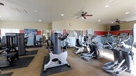 This single-floor fitness center offers a comprehensive workout environment designed to accommodate a range of exercise needs. The space is outfitted with a variety of cardio equipment, including stationary bikes, elliptical trainers, and additional aerobic machines, all arranged to allow comfortable movement between stations. Strength training equipment is also present, positioned along the perimeter to maximize usable floor space.

The room features a neutral color palette with warm beige walls accented by bold red horizontal bands, adding a sense of energy to the environment. Recessed ceiling lights provide consistent, even illumination throughout, while a ceiling fan promotes air circulation during workouts. Large mirrored panels line one wall, visually expanding the space and allowing users to monitor their form.

The flooring combines carpet in the main area with protective rubber mats beneath the equipment, balancing comfort and durability. Natural light filters in through windows, contributing to an open and inviting atmosphere. Overall, the space is practical, well-organized, and thoughtfully designed to support a productive fitness routine.
