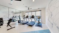 This contemporary fitness center is thoughtfully designed to provide a complete workout environment within a compact yet efficiently utilized single-level space. The room features a speckled epoxy floor that transitions into distinctive blue flooring near the entrance area, adding a bold visual contrast to the otherwise clean, white interior palette.

A full wall of floor-to-ceiling mirrors along one side visually expands the space while allowing users to monitor their form during exercise. Large, black-framed windows fitted with roller shades line the opposite wall, inviting ample natural light and offering views to the outdoors. A ceiling fan provides additional air circulation overhead, complementing the recessed lighting throughout.

The fitness area is equipped with multiple treadmills, elliptical machines, a stationary spin bike with an integrated display screen, and a dumbbell rack with an adjustable bench — catering to a range of cardio and strength training needs. A standout design element is the large-scale geometric mural of a running athlete rendered in fine lines and dots, adorning the rear wall and infusing the space with energy and artistic character.

A glass partition wall connects the fitness room to an adjacent area visible from the entrance, which features artwork and light wood flooring, suggesting a broader amenity suite. Overall, the space combines functional fitness infrastructure with modern, design-forward aesthetics.