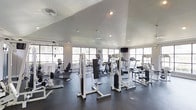 This fitness center in Garland is designed to support a comprehensive workout experience within a single-floor layout. The space features soaring vaulted ceilings that lend an open, airy atmosphere, complemented by recessed lighting and a ceiling fan that help maintain a comfortable environment during exercise. Large multi-pane windows line the perimeter walls, flooding the interior with abundant natural light and offering views of the surrounding greenery outside.

The rubber-floored workout area is well-organized and generously sized, accommodating an impressive array of equipment. Strength training is well represented with multiple cable and weight machines positioned throughout the space, providing options for a full-body resistance workout. The cardio section is equally well-appointed, featuring treadmills, elliptical trainers, and stationary bikes arranged near the windows to allow users to enjoy natural light while exercising. A wall-mounted television is also present, adding entertainment options for members. The clean white walls and neutral color palette contribute to a bright, functional atmosphere, making this fitness center a practical and inviting space for residents and users in the Garland area.
