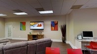 This single-floor space functions as a well-appointed lounge or common area, designed to offer both comfort and visual interest. The room features a drop-panel ceiling with recessed fluorescent lighting that provides bright, even illumination throughout the space. Durable wood-look flooring runs along the floor, complementing the neutral, warm-toned walls that serve as a backdrop for a curated selection of colorful abstract artwork, adding energy and personality to the room.

A large wall-mounted flat-screen television serves as a focal point, positioned above a rustic wooden media console that provides storage for equipment and accessories. Plush gray upholstered seating is arranged to face the screen, making the space ideal for group viewing or relaxed gatherings. Bold red accent chairs add a striking contrast and additional seating options.

A tall decorative vase with dried branches in a corner contributes an organic, artistic touch that ties the room's aesthetic together. The overall ambiance is casual yet thoughtfully styled, balancing functionality with a welcoming, lived-in comfort suited for lounging or socializing.