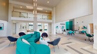 Located at 215 107th Street Circle East in Bradenton, Florida, this contemporary single-floor clubhouse offers a striking community gathering space defined by dramatic double-height ceilings and an open, flowing layout. The interior is anchored by an expansive main lobby and lounge area showcasing a bold, coastal-inspired color palette — vibrant teal and turquoise seating pieces are set against crisp white walls and light-toned herringbone and geometric-patterned tile flooring throughout. Floor-to-ceiling windows invite abundant natural light and frame views of the surrounding Florida landscape, including swaying palm trees. A glazed upper mezzanine level with glass railings adds architectural depth to the double-volume main space. The game and recreation area features a full-sized pool table beneath soaring ceilings, with an adjoining bar-height counter and wall-mounted television for social gatherings. Multiple distinct lounge seating zones are thoughtfully arranged throughout the space, including curved sectionals, swivel chairs, and a tufted emerald green banquette. A separate dining area accommodates a long communal table with upholstered chairs. A private meeting room offers a quiet retreat with a dark-topped table and lush outdoor views. A staffed leasing or service desk with computer stations completes the functional offerings of this well-appointed clubhouse.