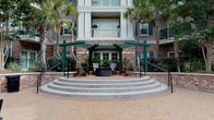 Located at 211 River Landing Drive on Daniel Island in Charleston, SC, Simmons Park presents an inviting and well-appointed community entrance defined by classic Lowcountry architectural character. The centerpiece of the arrival experience is a spacious brick porte-cochère, framed by substantial brick columns and accented with dark green architectural trim. Patterned brick pavers extend throughout the passage, leading residents and guests through an open-air corridor that flows seamlessly between the street and a lush interior courtyard. A second-level overlook with wrought iron railings adds vertical dimension and visual interest to the covered entry corridor, while seating areas with teak wood furniture and aqua-toned cushions offer a comfortable place to gather. The courtyard beyond the entrance is anchored by a charming pergola structure with Adirondack chairs arranged around a central fire pit, all framed by tall palm trees and manicured tropical plantings. A wide arched passageway frames views of the surrounding greenery and multi-story residential buildings. The branded entry doors, marked with the Simmons Park – Daniel Island identity, complete the polished and cohesive aesthetic of this thoughtfully designed community gateway.