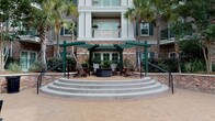 Located at 211 River Landing Drive on Daniel Island in Charleston, SC, Simmons Park presents an inviting and well-appointed community entrance defined by classic Lowcountry architectural character. The centerpiece of the arrival experience is a spacious brick porte-cochère, framed by substantial brick columns and accented with dark green architectural trim. Patterned brick pavers extend throughout the passage, leading residents and guests through an open-air corridor that flows seamlessly between the street and a lush interior courtyard. A second-level overlook with wrought iron railings adds vertical dimension and visual interest to the covered entry corridor, while seating areas with teak wood furniture and aqua-toned cushions offer a comfortable place to gather. The courtyard beyond the entrance is anchored by a charming pergola structure with Adirondack chairs arranged around a central fire pit, all framed by tall palm trees and manicured tropical plantings. A wide arched passageway frames views of the surrounding greenery and multi-story residential buildings. The branded entry doors, marked with the Simmons Park – Daniel Island identity, complete the polished and cohesive aesthetic of this thoughtfully designed community gateway.