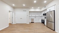 This single-floor unit presents a clean, contemporary design throughout. The open-concept layout connects the kitchen and main living area, creating a spacious, cohesive flow enhanced by recessed lighting and light-toned luxury vinyl plank flooring that runs consistently across the space.

The kitchen is well-appointed with crisp white shaker-style cabinetry featuring matte black hardware, extending from counter level to near the ceiling for generous storage capacity. A full suite of stainless steel appliances includes a French door refrigerator, electric range, built-in microwave, and dishwasher. White countertops and a stainless steel sink complete the functional workspace. A set of double doors adjacent to the kitchen provides additional storage or closet space.

The living area is open and versatile, with recessed lighting overhead and a window allowing natural light into the space. A dedicated bedroom area features neutral walls and the same consistent flooring.

The bathroom is finished with white hexagonal tile flooring, a white vanity with undermount sink, and a frameless sliding glass shower enclosure lined with large-format taupe ceramic wall tile, lending a polished, modern aesthetic. Overall, the unit presents a fresh, move-in-ready interior with consistent finishes throughout.