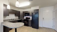This single-floor residence at Casanas Village at Frenchtown Square offers a thoughtfully designed open layout with modern finishes throughout. The kitchen is a standout feature, equipped with dark espresso cabinetry, granite-style countertops, a white subway tile backsplash, and a full suite of black appliances including a refrigerator, electric range, microwave, and dishwasher. A curved breakfast bar provides additional counter space and a casual dining option. Light wood-look flooring flows seamlessly throughout the living areas and bedrooms, creating a cohesive and contemporary feel. The living spaces are bright and airy, each outfitted with ceiling fans to ensure year-round comfort. Bedrooms are generously sized with ample natural light provided by window blinds. The bathrooms feature tile flooring, granite-topped dark vanities, and tub-shower combinations with white tile surrounds. A notable convenience is the in-unit stacked washer and dryer, adding practicality to daily living. Clean white walls and crisp trim work give the entire space a fresh, move-in-ready appearance, while the open-concept design between the kitchen and living area enhances the overall sense of space and flow.