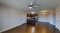 This single-floor space is designed with an open-concept layout that seamlessly connects the kitchen and living area, creating a spacious and functional environment. The kitchen is a standout feature, showcasing rich dark wood cabinetry that extends to the upper walls, granite-look countertops, and a full suite of stainless steel appliances including a refrigerator, microwave, and range. A central kitchen island provides additional prep and seating space, while pendant lighting fixtures hang above, adding a warm, focused glow to the area. The living space is anchored by warm, dark hardwood flooring that flows continuously throughout, lending a sense of cohesion and elegance. A ceiling fan with a built-in light fixture is centrally positioned, offering both comfort and illumination. Neutral-toned walls keep the space feeling bright and adaptable. A pendant light on the far end of the room hints at a defined dining area, while a standard entry door with clean white trim provides access to the unit. The overall ambiance is modern yet welcoming, with a practical layout well-suited for everyday living.