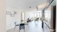 This contemporary one-bedroom, one-bathroom residence offers a well-appointed single-floor layout with a clean, modern aesthetic throughout. Light oak hardwood flooring and crisp white walls create a bright, cohesive atmosphere from room to room.

The open-concept living area is anchored by expansive floor-to-ceiling windows with black frames, flooding the space with natural light and offering sweeping city and mountain views. The living room comfortably accommodates a seating area and flows naturally into the dining space, which features a glass-top round table adjacent to the kitchen.

The kitchen is outfitted with a full suite of stainless steel appliances, including a range, dishwasher, and refrigerator, complemented by smoked glass upper cabinetry, light lower cabinetry, and a clean white stone backsplash and countertop.

The bedroom is a serene retreat, benefiting from the same floor-to-ceiling window wall and city views. It includes a double-door closet and ample room for a full bedroom suite.

The bathroom features large-format dark tile walls, a floating vanity with matte black fixtures, and a deep soaking tub with an overhead rain shower, delivering a spa-like finish. Every detail of this residence reflects refined, contemporary design.