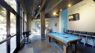This single-floor recreational space combines industrial aesthetics with vibrant, modern design to create a dynamic entertainment environment. The open-plan layout is anchored by a full-size pool table with a striking blue felt surface, accompanied by a wall-mounted cue rack and dartboard, making it a central hub for social activity. Exposed ductwork and track lighting run across the dark ceiling, casting warm, focused light throughout the space and reinforcing the industrial character. Concrete structural columns and polished concrete floors further complement this aesthetic, adding a sense of raw architectural texture. Bold sky-blue accent panels on the walls provide vivid contrast against the neutral tones, energizing the atmosphere. Tall windows along one side allow natural light to pour in, enhancing the open and airy feel of the space. A lounge area is visible further into the room, furnished with comfortable seating and complemented by wall art, offering a relaxed counterpoint to the more active recreation zone. Bar-height seating near the windows provides an additional social gathering spot. Together, these elements create a well-appointed, multi-purpose space that balances energetic recreation with comfortable lounging.