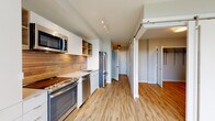 This single-floor residence showcases a contemporary design with a cohesive neutral palette and light wood-style flooring that extends throughout. The kitchen serves as a functional centerpiece, featuring a full suite of stainless steel appliances including a range, dishwasher, microwave, and refrigerator. White flat-front cabinetry is complemented by open wood shelving and a striking horizontal wood-plank backsplash that adds warmth and texture to the space. Light countertops and a single-basin sink complete the well-appointed layout.

The hallway connecting the main areas is lined with practical features, including a louvered storage door and utility access panels. Sliding barn-style doors appear throughout, offering a modern, space-conscious alternative to traditional hinged doors. The bedroom is generously proportioned, with multiple large windows that draw in natural light and frame treetop views, creating a bright and airy atmosphere. A walk-in closet with built-in shelving and rod storage is accessible through a sliding barn door.

The bathroom maintains the clean, minimalist aesthetic with neutral tile flooring, a soaking tub with tiled surround, a floating vanity, and recessed lighting overhead.