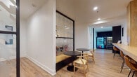 The Study Lounge is a thoughtfully designed, single-floor space that offers a variety of seating configurations to accommodate different study and collaboration needs. The overall aesthetic is clean and contemporary, characterized by light wood flooring, crisp white walls, and recessed lighting throughout.

The main corridor connects several distinct seating zones. A bar-height counter workspace, framed by warm wooden columns, provides individual seating with bar stools — ideal for focused, independent work. Adjacent to this area, small round black tables paired with natural wood wishbone chairs offer casual seating for one or two users.

A private enclosed booth features upholstered dark gray bench seating on both sides of a white rectangular table, creating an intimate and quiet setting for group study or focused work sessions. A glass partition with a geometric patterned film separates this nook from the main area, adding a decorative yet functional element.

A dedicated study room is equipped with a wall-mounted flat-screen television, a wood-topped rectangular table surrounded by red metal chairs, and a decorative potted plant, making it suitable for group collaboration or presentations. Artwork adds a personalized touch to the room.

Completing the space, a vending machine alcove at the end of the corridor provides convenient refreshment access for users throughout the lounge.