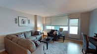 This single-floor space offers a thoughtfully designed and inviting living environment with a clean, modern aesthetic. The living room serves as the centerpiece, featuring warm parquet hardwood flooring that adds a classic touch to the space. Large windows fitted with roller blinds allow for excellent natural light control while brightening the room throughout the day. The space is furnished with a plush grey sofa and matching armchair arranged around a dark wood coffee table, all anchored by a soft area rug that adds warmth and texture underfoot. Subtle, neutral-toned walls provide a versatile backdrop that complements the contemporary furnishings. Tasteful décor, including framed wall art and coordinating accent lamps on side tables, contribute to a cohesive and polished interior. The textured ceiling adds a layer of architectural detail overhead. Overall, the space strikes a balance between comfort and style, making it a functional and welcoming environment suited for everyday living and relaxed entertaining.