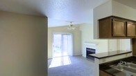 This single-floor space centers around a well-lit living area that immediately draws attention with its inviting atmosphere. A large sliding glass door fitted with vertical blinds fills the room with natural light while providing direct access to an outdoor area, offering a seamless connection to the exterior. The living area features wall-to-wall carpeting in a neutral tone that adds warmth underfoot, while the walls are painted in soft, muted yellow hues that contribute to the room's bright and airy feel.

A fireplace with a clean white mantle serves as a natural focal point along one wall, providing both aesthetic appeal and functional warmth. Overhead, a ceiling fan with an integrated light fixture ensures comfortable airflow and supplemental lighting throughout the space. The textured popcorn ceiling adds a classic, traditional character to the room.

Adjacent to the living area, the kitchen is partially visible and features dark wood cabinetry and a countertop that contrasts with the lighter tones of the living space. The open sightlines between the kitchen and living area create a cohesive, connected layout that enhances the overall sense of flow throughout this single-floor space.