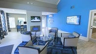 This single-floor space makes a strong visual impression through its thoughtfully designed lounge area, defined by a striking cobalt blue accent wall that anchors the room with vibrant energy. The seating arrangement centers around a sleek glass-top coffee table with a chrome frame, surrounded by neutral-toned armchairs with dark trim accents, all positioned atop a geometric patterned area rug in deep blues and grays. A contemporary wall-mounted fireplace serves as a refined focal point, complementing the room's modern aesthetic. Multiple flat-screen televisions are mounted throughout the space, enhancing both entertainment and informational display functionality. Decorative wall-mounted accents in white add artistic detail to the blue feature wall, while recessed lighting in the gray ceiling creates a clean, polished overhead finish. A mini-split HVAC unit ensures climate comfort. Adjacent areas visible through open sightlines suggest additional workspace or service zones, contributing to the overall versatility of the layout. Natural light filters in through windows, softening the bold color palette and brightening the interior. The overall ambiance is professional yet welcoming, well-suited for a reception, waiting, or community lounge setting.
