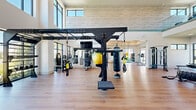 Located at 14300 Boggy Creek Rd in Orlando, FL, this professionally appointed fitness center offers a generously sized, single-floor exercise space designed to accommodate a wide variety of workout styles and training needs. The facility is characterized by soaring double-height ceilings in the main area, floor-to-ceiling windows, and clerestory windows above that flood the space with natural light and provide open views of the surrounding outdoor greenery.

The main workout floor features light wood-look flooring throughout, contributing to a bright and modern aesthetic. Equipment is thoughtfully arranged across the space and includes a TRX functional training rig with suspension straps and a heavy punching bag, selectorized strength machines, a cable multi-station system, kettlebells, battle ropes, a tire sled, medicine balls, and a treadmill, among other cardio and conditioning tools. Large wall-mounted mirrors in a dedicated section of the floor visually expand the space and support stretching or functional movement training.

A secondary area opens via a glass-paneled garage-style door directly to the outdoors, providing seamless indoor-outdoor access and additional ventilation. Ceiling fans are installed throughout to maintain air circulation. A water fountain station and storage cubbies for equipment are also present. The overall design strikes a balance between industrial utility and modern, clean-lined aesthetics, creating an inviting and high-functioning training environment.
