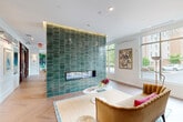 Located at 20013 Century Blvd in Germantown, MD, this single-floor space offers a thoughtfully designed interior that blends contemporary style with vibrant artistic elements. The expansive living room serves as the heart of the space, centered around a dramatic floor-to-ceiling fireplace clad in glossy emerald green stacked tile, which also features a mounted flat-screen TV. Herringbone-patterned hardwood flooring runs throughout, adding warmth and visual interest to the open layout. Large windows line the perimeter, flooding the interior with natural light and offering views of the surrounding greenery and streetscape. The living area transitions seamlessly into a game and dining zone, anchored by a striking marble-topped island, a billiard table, and a bold tropical mural that commands the far wall. Curated artwork, including botanical and wildlife prints, adds personality throughout. The kitchen is clean and modern, featuring white cabinetry, light countertops, a stainless steel sink, and an open display shelf styled with decorative accents. The entrance area features warm-toned seating, glass-paneled entry doors, and a welcoming lounge arrangement, completing this well-appointed and visually dynamic space.