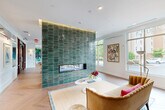 Located at 20013 Century Blvd in Germantown, MD, this single-floor space offers a thoughtfully designed interior that blends contemporary style with vibrant artistic elements. The expansive living room serves as the heart of the space, centered around a dramatic floor-to-ceiling fireplace clad in glossy emerald green stacked tile, which also features a mounted flat-screen TV. Herringbone-patterned hardwood flooring runs throughout, adding warmth and visual interest to the open layout. Large windows line the perimeter, flooding the interior with natural light and offering views of the surrounding greenery and streetscape. The living area transitions seamlessly into a game and dining zone, anchored by a striking marble-topped island, a billiard table, and a bold tropical mural that commands the far wall. Curated artwork, including botanical and wildlife prints, adds personality throughout. The kitchen is clean and modern, featuring white cabinetry, light countertops, a stainless steel sink, and an open display shelf styled with decorative accents. The entrance area features warm-toned seating, glass-paneled entry doors, and a welcoming lounge arrangement, completing this well-appointed and visually dynamic space.