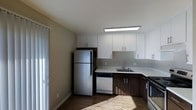 This single-floor space features a thoughtfully designed kitchen that serves as a central highlight. The kitchen showcases a clean, contemporary aesthetic with crisp white upper cabinetry contrasted by dark wood-toned lower cabinets, creating a visually appealing two-tone effect. Light-colored countertops provide ample workspace, complemented by a stainless steel sink and modern fixtures. Stainless steel appliances, including a full-sized refrigerator, are paired with a dishwasher and range, offering a complete suite of kitchen essentials. A flush-mounted ceiling light panel ensures consistent, even illumination throughout the kitchen area. The flooring features hardwood-style planks that add warmth and continuity to the space. Natural light filters in through a window, enhancing the bright and airy atmosphere. The neutral wall tones throughout create a versatile backdrop that complements the modern finishes. The overall design balances functionality with a clean, updated aesthetic, making it a practical and attractive space for everyday living.