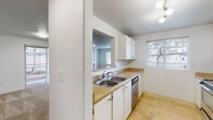 Located at 21163 NW Galice Ln in Portland, Oregon, this single-floor residence offers a well-organized layout designed for comfortable everyday living. The kitchen is a functional galley-style space finished with light wood flooring, white cabinetry, warm countertops, and a full suite of stainless steel appliances including a refrigerator, range, microwave, and dishwasher. A window above the sink brings in natural light, and a pass-through opening connects the kitchen to the main living area. The spacious living room features plush carpeting, neutral white walls, a baseboard heater, and an open feel that flows easily into the rest of the home. Bedrooms are carpeted and offer ample space, with at least one featuring a sliding-door closet with built-in shelving. The primary bathroom includes a dual-sink vanity with generous counter space and a separate room housing a soaking tub and toilet. A second bathroom provides accessibility-friendly features, including grab bars and a step-in shower. A dedicated laundry room includes a full-size washer and dryer with overhead wire shelving. The carpeted hallway connects all rooms seamlessly, and the clean, neutral palette throughout gives the space a bright, move-in-ready feel.