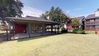 This single-story structure features a generously sized covered patio that serves as a prominent outdoor living area. The patio is defined by dark iron railings that line its perimeter, offering unobstructed views of the surrounding landscape while maintaining a clear boundary. The concrete flooring underfoot is clean and functional, and a recessed ceiling light provides overhead illumination. Large windows along the building's exterior wall allow natural light to flow between the interior and the covered outdoor space, creating a seamless visual connection.

Beyond the patio, a well-maintained, fenced grassy lawn extends outward, providing an open and airy feel. The iron fencing that encloses the lawn mirrors the railing style of the patio, lending a cohesive aesthetic to the outdoor environment. Mature shade trees are positioned throughout the grounds, adding greenery and natural character to the setting. Neatly trimmed shrubs are placed along the building's foundation and fence line, contributing to a tidy, landscaped appearance.

The surrounding community is visible from the grounds, with multi-story residential buildings featuring red and gray exteriors providing context and a sense of neighborhood. Under sunny skies, the outdoor spaces benefit from abundant natural light, creating a bright, welcoming atmosphere throughout the property.
