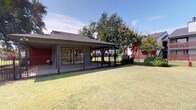 This single-story structure features a generously sized covered patio that serves as a prominent outdoor living area. The patio is defined by dark iron railings that line its perimeter, offering unobstructed views of the surrounding landscape while maintaining a clear boundary. The concrete flooring underfoot is clean and functional, and a recessed ceiling light provides overhead illumination. Large windows along the building's exterior wall allow natural light to flow between the interior and the covered outdoor space, creating a seamless visual connection.

Beyond the patio, a well-maintained, fenced grassy lawn extends outward, providing an open and airy feel. The iron fencing that encloses the lawn mirrors the railing style of the patio, lending a cohesive aesthetic to the outdoor environment. Mature shade trees are positioned throughout the grounds, adding greenery and natural character to the setting. Neatly trimmed shrubs are placed along the building's foundation and fence line, contributing to a tidy, landscaped appearance.

The surrounding community is visible from the grounds, with multi-story residential buildings featuring red and gray exteriors providing context and a sense of neighborhood. Under sunny skies, the outdoor spaces benefit from abundant natural light, creating a bright, welcoming atmosphere throughout the property.