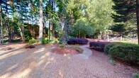 Located in Smyrna, Georgia, this single-level outdoor amenity space at Vinings Trail SE offers a well-maintained and naturally shaded environment designed for community recreation and relaxation. The grounds are defined by a rich canopy of mature trees — including towering hardwoods and a distinctive ornamental evergreen — that provide abundant shade throughout the space. Mulched planting beds frame a gently curving concrete pathway that winds through the landscaped grounds, guiding visitors through the various areas of the property. Decorative shrubs with deep purple and green foliage add layers of color and texture to the naturalistic setting. A dedicated picnic and grilling area features a wooden picnic table alongside a pedestal-mounted charcoal grill, set against a backdrop of dense woodland and a privacy fence — ideal for outdoor gatherings. Crushed gravel ground cover throughout contributes to the rustic, park-like character of the space. Toward one end of the property, a covered pavilion with a hip roof and ceiling fans is situated adjacent to a fenced community pool area, enclosed by ornamental black metal fencing. The combination of mature landscaping, recreational amenities, and natural surroundings makes this outdoor space an inviting and functional extension of the community.