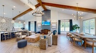 Located at 24 Winnow Walk in Wilmington, NC, this thoughtfully designed single-story community clubhouse offers a generous, open-concept layout filled with natural light and upscale finishes throughout. The centerpiece of the space is a dramatic vaulted ceiling adorned with rich, exposed wood beams, anchored by a striking blue paneled accent wall with a mounted flat-screen TV and flanked by an elegant candle-style chandelier. Herringbone-patterned light wood flooring flows seamlessly across the entire space, lending warmth and visual continuity. The main gathering area features comfortable lounge seating arranged around live-edge wood coffee tables, complemented by a large formal dining table with modern upholstered chairs beneath a contemporary chandelier. Abundant windows dressed with navy drapery invite natural light and offer views of the surrounding greenery. A billiards table adds a recreational focal point in an adjoining zone, while additional casual round tables and chairs provide flexible seating. The well-appointed cabinetry area features light blue-gray lower cabinets, white quartz countertops, a black matte faucet, and a bold geometric-patterned tile accent wall. Curated artwork is displayed throughout, reinforcing the space's coastal-contemporary aesthetic. Direct access to an outdoor pool area further enhances this clubhouse's appeal as a premier community gathering destination.