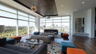 This sophisticated single-floor space makes a striking first impression with its expansive floor-to-ceiling glass curtain walls that flood the interior with natural light and frame breathtaking panoramic views of the surrounding cityscape and greenery beyond. The soaring ceiling height is accentuated by a rich wood slat ceiling detail that adds warmth and architectural depth, complemented by a statement sputnik chandelier and a secondary pendant light that provide both ambient and accent illumination. The open-plan lounge area is thoughtfully arranged with a curated mix of contemporary seating, including modular sofas, sleek ottomans in bold teal and orange tones, and minimalist side tables that contribute to the space's vibrant, mid-century modern aesthetic. A multicolored geometric area rug anchors the central seating arrangement, injecting playful energy into the design. Dark hardwood flooring runs throughout, providing an elegant contrast to the bright, airy atmosphere created by the glass walls. Multiple distinct seating clusters allow for both intimate conversation and larger group gatherings. The overall ambiance is one of elevated comfort and modern sophistication, with a dynamic interplay of natural light, urban views, and carefully selected design elements that make this space as functional as it is visually compelling.