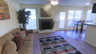 This single-story space centers around a welcoming living area designed for comfort and functionality. The room's focal point is a classic brick fireplace with a raised hearth, adding warmth and a traditional character to the space. A ceiling fan light fixture overhead provides both ambient lighting and air circulation throughout the room. Multiple windows along the walls invite generous amounts of natural light, creating a bright and airy atmosphere, while a glass-panel exterior door enhances the connection between the interior and the outdoors. The flooring transitions between carpet and hard surface areas, offering both comfort and practicality. A large area rug with a bold geometric circle pattern in earth tones and blues anchors the seating area, adding a modern, decorative contrast to the room's more traditional elements. Decorative wall art above the fireplace and a tall indoor plant in the corner contribute to the room's lived-in, homey ambiance. Overall, the living space balances classic architectural details with everyday comfort.