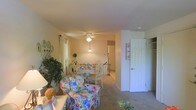 This two-floor residential space offers a welcoming and cozy atmosphere throughout. The upper level showcases an open layout that combines a casual sitting area and a dining space, creating a versatile and functional living environment. The sitting area features plush, floral-upholstered seating that adds a warm, homey character to the space, complemented by lush indoor plants that bring a touch of greenery indoors. The dining area is centered around a white wooden table with matching chairs, ideal for gatherings and everyday meals. A ceiling fan with a light fixture provides both comfort and illumination, while multiple windows dressed with curtains invite ample natural light, enhancing the bright and airy feel of the room. Neutral-toned carpet runs throughout, adding warmth underfoot. A staircase with a decorative railing connects the two floors, offering easy access between levels. A paneled interior door leads to additional rooms, suggesting further living or storage space beyond. The overall ambiance is warm and residential, with classic design elements that create a comfortable and lived-in environment suitable for everyday living.