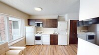 This single-floor unit is designed for efficient, comfortable living, with a thoughtful layout that maximizes functionality throughout. The main living area features warm-toned laminate flooring, neutral walls, and multiple windows fitted with horizontal blinds that invite natural light while maintaining privacy. A wall-mounted air conditioning unit provides climate control, and a wall-mounted television adds a modern convenience. The space accommodates a tufted sofa, coffee table, and a small dining set, making it suitable for everyday living and casual entertaining. A large built-in wardrobe provides ample storage within the living space. The kitchen runs along one wall and is equipped with a full-size refrigerator, electric range, dishwasher, and dark-stained cabinetry providing generous storage. A stacked washer and dryer unit is integrated directly into the kitchen area, offering convenient in-unit laundry. A wall-mounted microwave completes the kitchen setup. The bathroom features light wood-style flooring, a single-basin vanity with a modern cabinet, a large mirror, and a walk-in shower stall with a curtain rod. Textured ceilings and consistent flooring throughout tie the space together cohesively.
