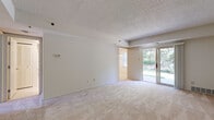 This single-floor unit offers a straightforward and functional layout designed to maximize everyday comfort. The living area serves as the central gathering space, featuring neutral-toned carpeting, a textured ceiling, and a large sliding glass door that floods the room with natural light while providing direct views of the lush greenery outside. A dining or secondary living area is visible beyond, connected seamlessly to the main space.

The kitchen is equipped with warm-toned wood cabinetry that extends to the upper walls, offering generous storage capacity. Dark countertops provide a practical contrast, and the space includes a full suite of white appliances — a refrigerator, range, microwave, and dishwasher. Light tile flooring completes the room's clean, utilitarian aesthetic.

The bedrooms are carpeted and finished with neutral walls, each featuring louvered bi-fold closet doors that provide organized storage. One bedroom benefits from a window, allowing natural light to filter in. The bathroom is finished in crisp white tile flooring and includes a full soaking tub with an overhead shower, a toilet, and a louvered storage closet, maintaining a clean and functional design throughout.