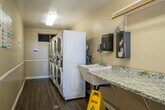 Located at 1939 Wallace Avenue in Costa Mesa, California, this shared laundry facility offers a practical and well-organized space for residents. The room is laid out in a narrow, elongated floor plan that makes efficient use of its compact footprint.

Along one wall, a row of stacked front-loading washers and dryers provides ample laundry capacity. The machines are modern, white units equipped with digital card-payment systems, offering a convenient and cashless experience. A small utility sink is also present, adding to the room's functionality.

Opposite the machines, a long granite-topped folding counter runs the length of the wall, providing generous surface area for sorting and folding laundry. Above the counter, a wall-mounted shelf with a hanging rod offers additional organizational space. Informational notices and usage instructions are posted on the walls for residents' reference.

The room features durable wood-look plank flooring and is finished with neutral beige walls and white wainscoting trim, giving it a clean and tidy appearance. Overhead fluorescent lighting ensures the space is well-lit. Natural light filters in through windows and a glass-panel entry door, which opens to an outdoor courtyard, contributing to a bright and airy atmosphere.
