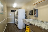 Located at 1939 Wallace Avenue in Costa Mesa, California, this shared laundry facility offers a practical and well-organized space for residents. The room is laid out in a narrow, elongated floor plan that makes efficient use of its compact footprint.

Along one wall, a row of stacked front-loading washers and dryers provides ample laundry capacity. The machines are modern, white units equipped with digital card-payment systems, offering a convenient and cashless experience. A small utility sink is also present, adding to the room's functionality.

Opposite the machines, a long granite-topped folding counter runs the length of the wall, providing generous surface area for sorting and folding laundry. Above the counter, a wall-mounted shelf with a hanging rod offers additional organizational space. Informational notices and usage instructions are posted on the walls for residents' reference.

The room features durable wood-look plank flooring and is finished with neutral beige walls and white wainscoting trim, giving it a clean and tidy appearance. Overhead fluorescent lighting ensures the space is well-lit. Natural light filters in through windows and a glass-panel entry door, which opens to an outdoor courtyard, contributing to a bright and airy atmosphere.