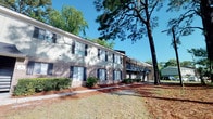 Situated at 12012 Middleground Rd in Savannah, Georgia, this single-floor unit is part of a well-established two-story residential building that blends classic brick construction with a welcoming, tree-lined environment. The exterior features a traditional brick façade accented by navy blue shutters and framed by towering pine trees that provide natural shade and a sense of seclusion. Lush shrubbery lines the building's perimeter, contributing to the overall greenery and curb appeal of the property. The building includes exterior staircase access and an open-air upper corridor, characteristic of a well-maintained multi-unit residential structure. The surrounding grounds are spacious and open, offering a relaxed, park-like atmosphere that reflects the quiet, suburban character of the Savannah area. The mature landscaping and tree canopy create a naturally shaded environment, particularly appealing in the warm Georgia climate. This property offers a comfortable setting within reach of the amenities and services of Savannah, making it a practical and appealing residential option in the 31419 zip code.
