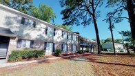 Situated at 12012 Middleground Rd in Savannah, Georgia, this single-floor unit is part of a well-established two-story residential building that blends classic brick construction with a welcoming, tree-lined environment. The exterior features a traditional brick façade accented by navy blue shutters and framed by towering pine trees that provide natural shade and a sense of seclusion. Lush shrubbery lines the building's perimeter, contributing to the overall greenery and curb appeal of the property. The building includes exterior staircase access and an open-air upper corridor, characteristic of a well-maintained multi-unit residential structure. The surrounding grounds are spacious and open, offering a relaxed, park-like atmosphere that reflects the quiet, suburban character of the Savannah area. The mature landscaping and tree canopy create a naturally shaded environment, particularly appealing in the warm Georgia climate. This property offers a comfortable setting within reach of the amenities and services of Savannah, making it a practical and appealing residential option in the 31419 zip code.