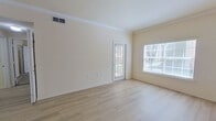 This single-story, one-bedroom, one-bathroom unit offers a thoughtfully organized layout that maximizes its compact footprint. The living room serves as the heart of the space, finished with light wood-style flooring and accented by crown molding along the ceiling. Large windows and a glass-paneled exterior door flood the area with natural light while providing outdoor views, creating an open and airy atmosphere. The entry door features a classic six-panel design, adding a traditional architectural touch.

The kitchen is positioned adjacent to the living and dining area and features light wood cabinetry, white appliances including a range, microwave, and refrigerator, and tile flooring. A peninsula-style counter provides a natural boundary between the kitchen and the adjoining space, with pendant lighting overhead adding functional illumination.

The bedroom is a generously proportioned room with plush carpet flooring, a ceiling fan with light fixture, and a window that admits ample natural light. A dedicated walk-in closet off the bedroom offers wire shelving on multiple levels, providing practical and organized storage.

The bathroom includes a single-sink vanity with white cabinetry, a large mirror, tile flooring, and a full tub-shower combination. Neutral, warm beige tones are carried consistently throughout the unit, contributing to a clean and cohesive aesthetic.