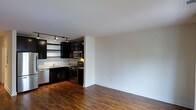 This single-floor space offers a clean, open-concept layout that seamlessly connects the living area and kitchen, creating a sense of spaciousness and ease of movement. The kitchen is outfitted with sleek, dark espresso cabinetry that extends to the upper walls, providing generous storage while delivering a bold, contemporary aesthetic. Light stone countertops contrast elegantly with the dark cabinetry, and the space is equipped with stainless steel appliances, including a gas range, over-the-range microwave, and dishwasher. A track lighting system mounted on the ceiling ensures the kitchen is well-lit and functional. The open living area flows naturally from the kitchen, bathed in a neutral white palette that keeps the space feeling bright and airy. Rich, warm-toned hardwood flooring runs throughout, anchoring the space with a sense of warmth and continuity. The minimalist white walls offer a versatile canvas, allowing the architectural details and finishes to take center stage. The overall ambiance is modern and refined, balancing functionality with a sophisticated design sensibility.