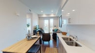 This single-floor residence features a thoughtfully designed open-concept layout with dark hardwood flooring throughout and a clean, contemporary aesthetic. The living area is anchored by large French doors leading to a private balcony, allowing abundant natural light and offering broad outdoor views of the surrounding cityscape. A tufted leather Chesterfield sofa and organic wood accent tables create a warm, inviting atmosphere in the living space.

The kitchen is well-appointed with two-tone cabinetry — white upper cabinets and light wood-grain lower cabinets — paired with white quartz countertops and a full suite of stainless steel appliances, including a gas range, over-the-range microwave, and refrigerator. A built-in breakfast bar provides additional workspace and casual seating.

The bedroom is bright and serene, featuring a large window with sweeping views and ample space for a full-sized bed and furnishings. The bathroom offers a generous vanity with dual sinks and a soaking tub, finished with wood-look flooring that maintains a cohesive feel with the rest of the unit. A conveniently located in-unit washer is tucked behind louvered doors in the hallway, adding practical functionality to the space.