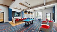 Located at Waterfront Crossing in Greenacres, Florida, this single-story community amenity space delivers a dynamic and entertainment-focused environment designed for social gatherings and recreation. The interior is defined by bold cobalt blue grasscloth accent walls, vivid multicolored striped carpeting, and rich wood-toned ceiling beams and wall panels that add warmth to the contemporary design.

The cinema lounge area features a curated gallery of iconic movie posters — including classic films such as E.T., Kill Bill, and Goodfellas — displayed against a deep blue accent wall. Plush swivel chairs with red accent pillows, a round coffee table, and a large wall-mounted flat-screen television create an intimate screening atmosphere, complemented by a popcorn cart for a true theater experience.

Transitioning into the main recreation area, a full-size billiards table takes center stage beneath a pair of striking oversized pendant lights. A wet bar with a kitchen counter, bar seating with red stools, and additional dining tables provide ample space for socializing. A foosball table and a bold Toy Story-themed collage mural energize the game zone. Large windows along the perimeter invite natural light and offer views of the outdoor surroundings, while hardwood-style flooring anchors the space with a clean, polished finish.