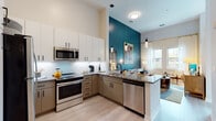This single-floor residence showcases a thoughtfully designed open-concept layout with soaring ceilings that lend a sense of spaciousness throughout. The kitchen is a functional highlight, featuring two-tone cabinetry with white upper cabinets and taupe lower cabinets, granite countertops, a subway tile backsplash, and a full suite of stainless steel appliances including a refrigerator, range, microwave, and dishwasher. A peninsula with seating for two is illuminated by sleek pendant lighting, making it ideal for casual dining. The kitchen flows seamlessly into the living area, which is distinguished by a dramatic teal accent wall rising to the tall ceiling, providing a striking backdrop for artwork and decor. Large windows invite abundant natural light and offer views of surrounding greenery. The bedroom is a tranquil retreat featuring generous closet space and large windows framing lush outdoor scenery. The bathroom is finished with marble-look countertops, subway-tiled tub surround, and matte black fixtures throughout. Light wood-style flooring unifies the entire residence, contributing to its clean, contemporary aesthetic.