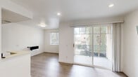 This single-floor unit is designed with a clean, modern aesthetic, featuring crisp white walls, recessed lighting, and light-toned wood-style flooring that runs consistently throughout the space. The living area is open and airy, benefiting from generous natural light provided by large windows fitted with horizontal blinds and a full-width sliding glass door that opens onto a private balcony with views of surrounding greenery and neighboring structures. The layout flows naturally from the living area into the kitchen, which is equipped with dark cabinetry, a mosaic tile backsplash, light countertops, stainless steel appliances including a gas range, microwave, and refrigerator, and a peninsula that provides additional prep space. Multiple bedrooms offer versatile use, each featuring the same light flooring and white walls. One bedroom includes a mirrored sliding closet door, while others present a clean, flexible canvas. Two full bathrooms are each fitted with a tub-shower combination enclosed by sliding glass doors with chrome framing. The overall ambiance of the unit is bright, neutral, and move-in ready, with a consistent design language that makes the space feel cohesive and well-maintained throughout.