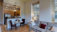 Located at 300 Blackwell St in the heart of Durham, NC, this single-floor unit offers a thoughtfully designed open-concept layout that seamlessly blends living, dining, and kitchen areas. The living space is anchored by a generously sized sofa and accented by a large, floor-to-ceiling window that draws in abundant natural light, highlighting the warm hardwood flooring throughout. The impressive ceiling height adds a sense of openness and airiness rarely found in urban residences. A compact dining area with a glass-top table and leather chairs sits adjacent to the kitchen, creating an inviting space for casual meals. The kitchen features warm wood cabinetry, a built-in microwave, and a convenient pass-through counter that connects it to the living and dining area, enhancing the open feel. Pendant lighting above the kitchen area adds a stylish, modern touch. The exposed structural elements visible through the large window hint at the building's industrial heritage, blending historic character with contemporary finishes. The overall ambiance is bright, clean, and modern, making this a comfortable and functional urban living space in one of Durham's most dynamic neighborhoods.