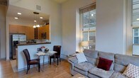 Located at 300 Blackwell St in the heart of Durham, NC, this single-floor unit offers a thoughtfully designed open-concept layout that seamlessly blends living, dining, and kitchen areas. The living space is anchored by a generously sized sofa and accented by a large, floor-to-ceiling window that draws in abundant natural light, highlighting the warm hardwood flooring throughout. The impressive ceiling height adds a sense of openness and airiness rarely found in urban residences. A compact dining area with a glass-top table and leather chairs sits adjacent to the kitchen, creating an inviting space for casual meals. The kitchen features warm wood cabinetry, a built-in microwave, and a convenient pass-through counter that connects it to the living and dining area, enhancing the open feel. Pendant lighting above the kitchen area adds a stylish, modern touch. The exposed structural elements visible through the large window hint at the building's industrial heritage, blending historic character with contemporary finishes. The overall ambiance is bright, clean, and modern, making this a comfortable and functional urban living space in one of Durham's most dynamic neighborhoods.