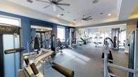 This fitness center is designed to support a comprehensive workout experience within a spacious, single-level layout. The room is distinguished by bold navy blue accent walls that contrast with crisp white ceilings, creating an energetic yet focused atmosphere. Two large ceiling fans are mounted overhead, ensuring consistent air circulation throughout the space, while recessed lighting provides even illumination across the entire floor.

The facility is divided into functional zones. The strength training area lines one side of the room and features a variety of resistance machines and free weight equipment, including cable machines, leg press stations, and benches. Full-length mirrors are strategically positioned along the walls, enhancing the sense of openness while allowing users to monitor their form. The cardio section, situated toward the opposite end, houses a row of treadmills and stair climbers, bathed in natural light from large windows along the perimeter.

The flooring consists of durable, striped carpeting suited for a fitness environment. The overall ambiance is clean, modern, and purposeful, making this fitness center a well-appointed space for a full range of exercise activities.