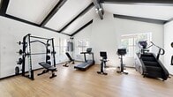 The Missions at Back Bay Amenity Gym is a 540-square-foot fitness facility located in Costa Mesa, CA, designed to provide residents with a well-equipped and architecturally appealing workout environment. The single-room gym is defined by its dramatic vaulted ceilings accented with bold, dark exposed beams set against crisp white shiplap, creating a bright and airy atmosphere that feels open and inviting. Natural light floods the space through multiple windows that frame views of the surrounding greenery and outdoor pool area, further enhancing the uplifting ambiance. The light wood-tone flooring complements the clean, modern aesthetic throughout. The gym is outfitted with a comprehensive selection of equipment to support a full range of fitness goals. Cardio offerings include a treadmill, upright stationary bike, stair climber, and elliptical machine. For strength training, the facility features a Smith machine with an adjustable bench, a dual cable functional trainer with pull-up bar, and a rack of dumbbells. A ceiling fan and wall-mounted air conditioning unit ensure comfort during intense workouts. With two access points and windows opening to outdoor views, this thoughtfully designed amenity gym delivers both functionality and a pleasant workout experience for residents.
