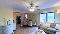 This single-floor residence offers a comfortable and cohesive living environment defined by an open-concept layout that seamlessly connects the main living areas. The living room is anchored by a plush sofa, a coffee table with a decorative tray, and a cozy area rug, all bathed in natural light streaming through a large window that looks out onto lush greenery. A ceiling fan with a light fixture overhead adds both function and a classic touch to the space.

Adjacent to the living room, the dining area features a round table with upholstered chairs and a decorative metal wall accent, contributing to the room's warm, welcoming character. Warm-toned walls in a soft terracotta hue complement the rich hardwood-style flooring that flows throughout the main living spaces.

The kitchen is visible through an arched passageway and is equipped with wooden cabinetry, black appliances, and a breakfast bar that provides both additional prep space and a casual seating option. The overall ambiance of the home is cozy and lived-in, with warm color tones and tasteful decorative accents that create an inviting atmosphere throughout.