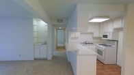 This single-story space presents a practical and straightforward layout designed for everyday living. The kitchen is positioned along one side of the main living area and features white cabinetry, white appliances including a gas range and microwave, and a dual-basin sink set into a continuous light-colored countertop. An angled peninsula extends outward, creating a natural division between the kitchen and the adjacent carpeted living area while also providing additional workspace. An overhead skylight or flush light fixture illuminates the kitchen, keeping the space bright and functional. A secondary wet bar or utility area is visible to the left, equipped with its own sink, white cabinetry, and open shelving, offering additional storage and convenience. A long central hallway extends toward the rear of the unit, lined with doors that lead to additional rooms. The flooring transitions from hardwood or laminate in the kitchen to carpet in the living and hallway areas. The overall color palette is neutral throughout, with white walls, white trim, and light-toned surfaces contributing to a clean, understated aesthetic. Ceiling vents are visible, indicating a central air system is in place.